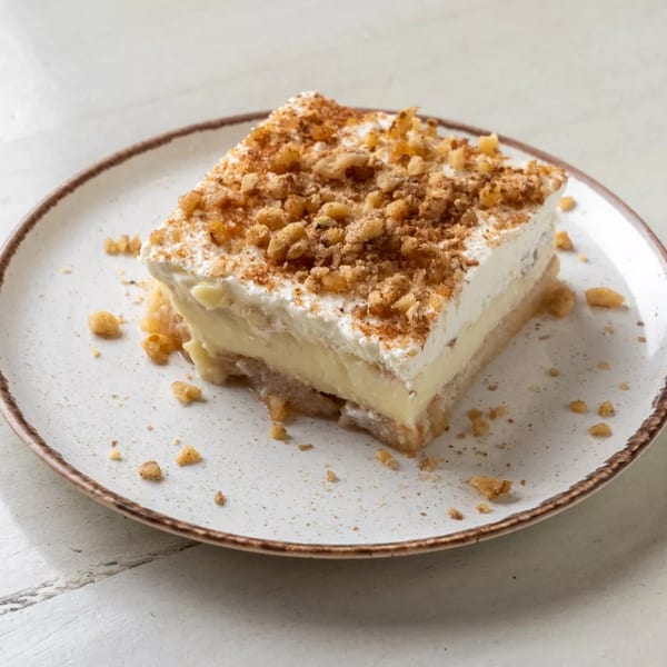 A square slice of dessert with a white cream topping and crushed nuts on a speckled white plate.