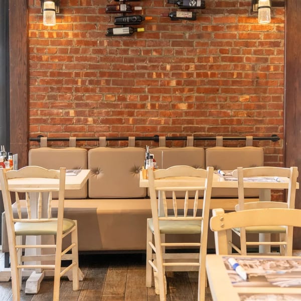 Restaurant interior with brick wall, wine bottles, booth seating, and light wooden tables and chairs.