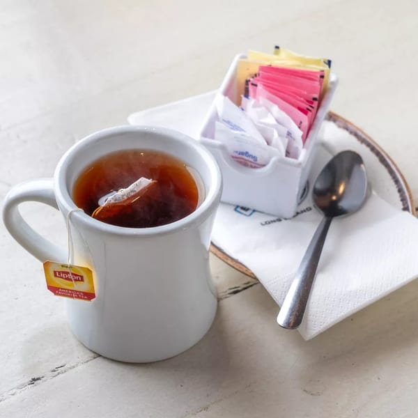 A white mug of Lipton tea with a tea bag, sugar packets, and spoon on a white table.