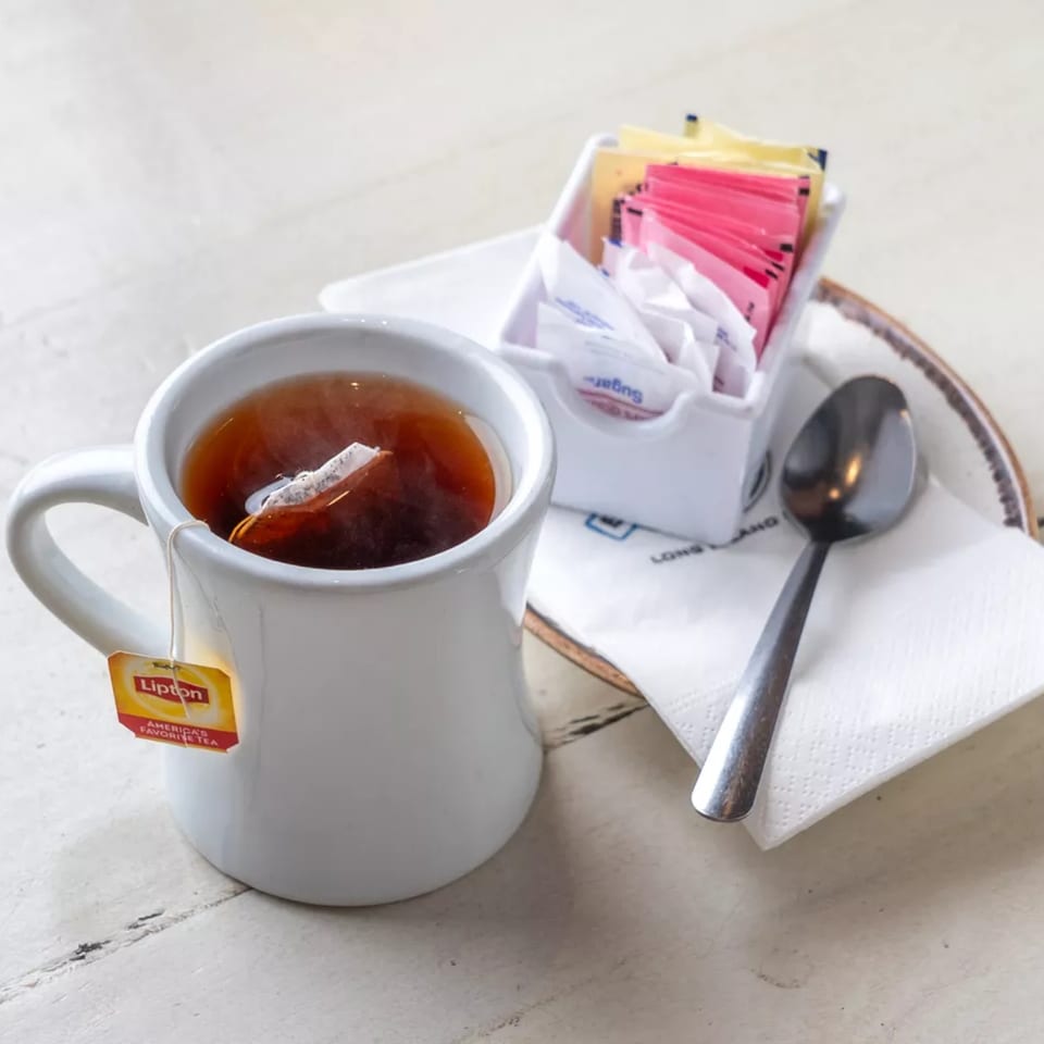 A white mug of Lipton tea with a tea bag, sugar packets, and spoon on a white table.
