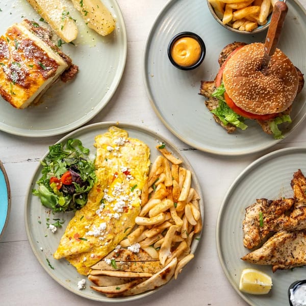 Overhead view of four plates with various Greek dishes, including moussaka, omelet, burger, and grilled chicken.